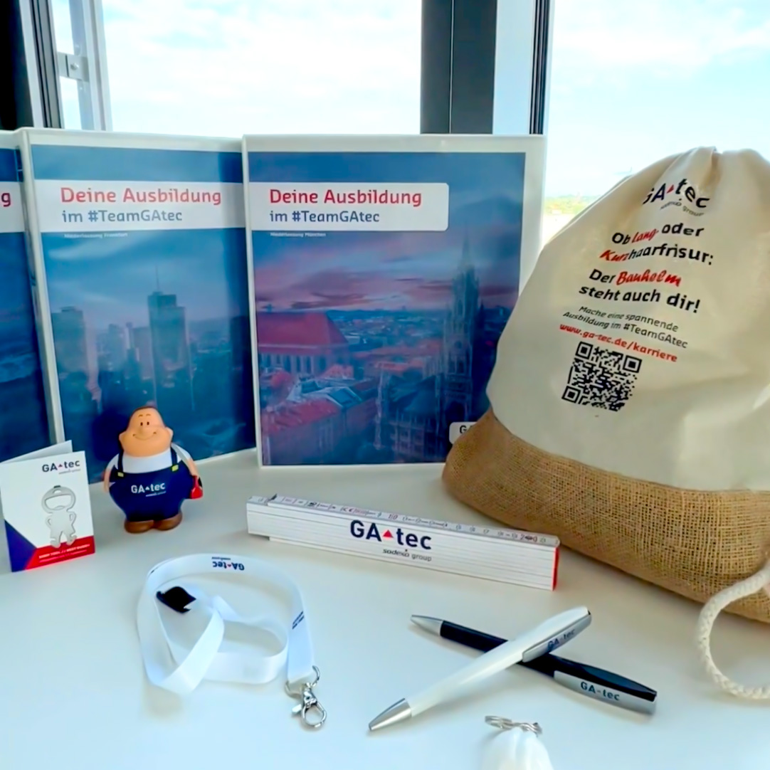 Werbematerialien von GA-tec zur Ausbildung im #TeamGAtec auf einem Tisch: Drei Ordner mit der Aufschrift ‚Deine Ausbildung im #TeamGAtec‘, ein beiger Jutebeutel mit roter und blauer Schrift sowie QR-Code, ein Maßstab mit GA-tec Logo, zwei Kugelschreiber, ein weißes Lanyard, eine kleine Figur mit GA-tec Logo, ein Schlüsselanhänger u. v. m.